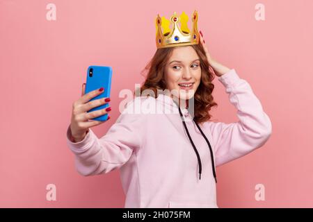 Porträt eines lächelnden, lockigen Teenagers im Hoodie mit goldener Krone, das auf der Smartphone-Kamera posiert und Selfie macht. Innenaufnahme des Studios isoliert auf rosa Hintergrund Stockfoto