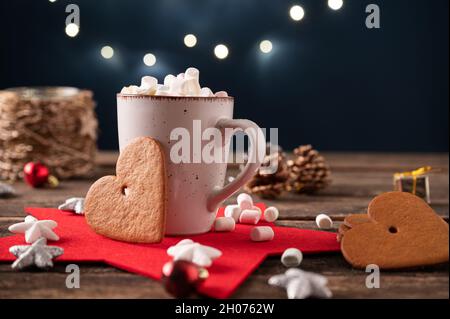 Heißer Kakao mit kleinen Marschwalben in einer rustikalen Tasse auf einem Holzschreibtisch voller weihnachtsdekorationen und großen herzförmigen Keksen. Stockfoto