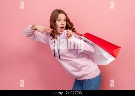 Porträt eines überraschten lockigen Teenagers im Hoodie mit offenem Mund und Zeigefinger auf Papiertüten hinter dem Rücken, großem Rabatt, Verkauf. Innenaufnahme des Studios isoliert auf rosa Hintergrund Stockfoto