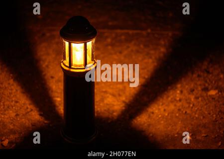 Lampe oder Licht in der Nacht in einem Park mit Schatten in einem kreisförmigen Muster. Das Licht beleuchtet einen steinigen Weg. Off-Center-Ansicht der Lampe mit Spinnennetz. Stockfoto