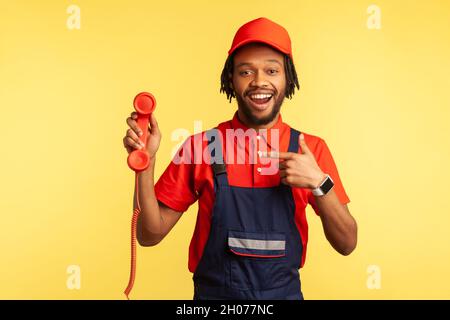 Zufriedener bärtiger Arbeiter in Uniform mit rotem Festnetztelefon, zeigt auf Hörer, wartet auf Aufträge, ist bereit zur Arbeit. Innenaufnahme des Studios isoliert auf gelbem Hintergrund. Stockfoto