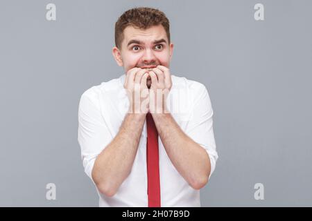 Porträt eines jungen nervösen Mannes in weißem Hemd und Krawatte stehen, seine Nägel bissen und suchen mit nervösen Sorgen Gesicht. Indoor isoliert auf grauem Hintergrund. Stockfoto