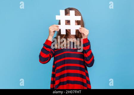 Neugierige Frau, die einen gestreiften Pullover im lässigen Stil trägt und durch ein großes weißes Hashtag-Symbol mit neugierigem Auge und interessanten Webinhalten auf die Kamera blickt. Innenaufnahme des Studios isoliert auf blauem Hintergrund. Stockfoto