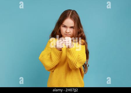 Wütendes kleines Mädchen, das mit erhobenen Fäusten steht, bereit ist zu kämpfen, mit ernsthaftem Ausdruck aussieht und einen gelben Pullover im lässigen Stil trägt. Innenaufnahme des Studios isoliert auf blauem Hintergrund. Stockfoto