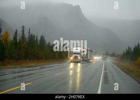 Ein LKW mit einem vorgefertigten Wohnmobil bei schlechtem Wetter, wo die Sicht aufgrund von Nebel und Regen eingeschränkt ist. Helferautos warnen den Verkehr zum w Stockfoto