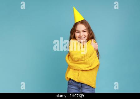 Kleines Mädchen im Partykegel, umarmt sich, feiert ihren Geburtstag, schaut lächelnd auf die Kamera, trägt gelben Pullover im lässigen Stil. Innenaufnahme des Studios isoliert auf blauem Hintergrund. Stockfoto