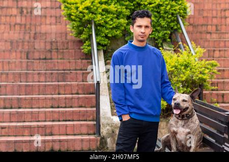 Mann in blauen Kleidern posiert neben seinem Haustier Pitbull mit orangen Treppen im Hintergrund. Stockfoto