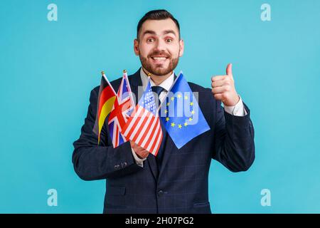 Porträt eines glücklichen bärtigen Geschäftsmannes in einem offiziellen Anzug mit Flaggen von Deutschland, den USA, Großbritannien und der Europäischen Union, der den Daumen nach oben zeigt. Innenaufnahme des Studios isoliert auf blauem Hintergrund. Stockfoto