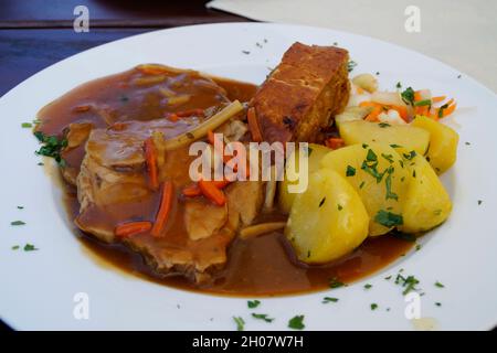 Köstliches österreichisches Gericht, bestehend aus Schweinebraten, Kartoffeln und einem Stück Kuchen, übergossen mit einer großen Portion Fleischsoße Stockfoto