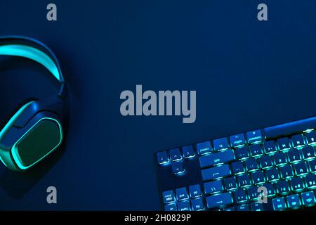 Draufsicht auf cyanfarbene Computertastatur und Kopfhörer mit Hintergrundbeleuchtung vor dunklem Hintergrund. Professionelle Computerspiele, eSport und so weiter Stockfoto