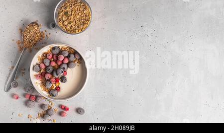 Schüssel mit hausgemachtem Joghurt mit Müsli und gefrorenen Beeren bedeckt mit Reif auf grauem Hintergrund, Draufsicht mit Kopierraum Stockfoto