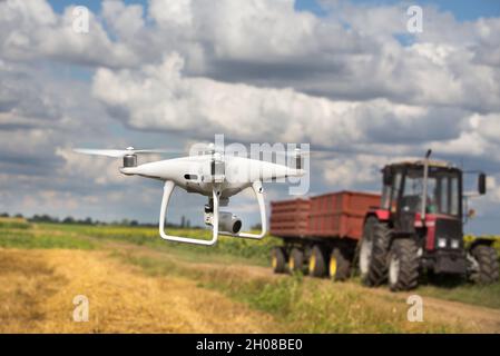 Drohne fliegt im Frühsommer vor dem Traktor mit Anhänger im Feld. Technologische Innovationen in der Landwirtschaft Stockfoto