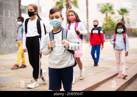 Schuljunge in medizinischer Maske in der Nähe der Schule, Kinder auf dem Hintergrund Stockfoto