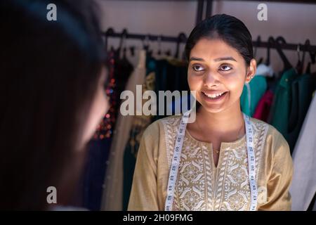 Junge glückliche indische Frau, Schneiderin Näherin, lächelnde Modedesignerin im Gespräch mit Kunden oder Kunden, stehend mit Maßband in Kleidungswerkstatt, a Stockfoto