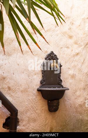 Alter kunstvoller Messing-Wasserhahn und -Becken, die an einer weißen verputzten Wand in einem Garten befestigt sind Stockfoto