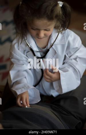 Ein kleines Mädchen im Mantel eines Arztes untersucht ihren Großvater mit einem Stethoskop. Kinderspiele Stockfoto