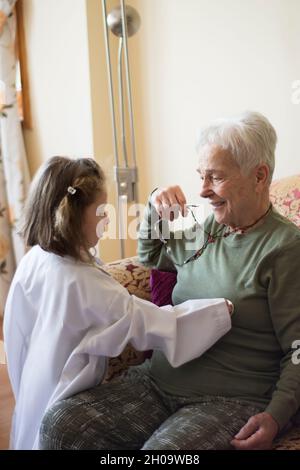 Ein kleines Mädchen im Mantel eines Arztes untersucht ihre Großmutter mit einem Stethoskop. Kinderspiele. Selektives Focu Girl Stockfoto