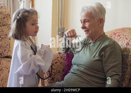 Ein kleines Mädchen im Mantel eines Arztes untersucht ihre Großmutter mit einem Stethoskop. Kinderspiele. Selektives Focu Girl Stockfoto