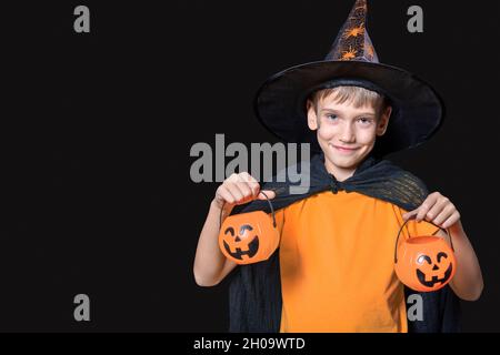 Halloween Kinder. Junge in Zaubererhut und orangefarbenem T-Shirt mit Kürbis-förmigen Eimern von Süßigkeiten, die auf schwarzem Hintergrund isoliert sind. Bereit für die ho Stockfoto