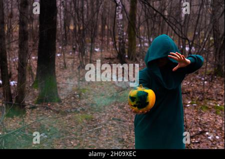 Eine gruselige Hexe hält einen dampfenden Kürbis in einen tiefen Wald. Jack o Laterne mit grünem Rauch für halloween Stockfoto