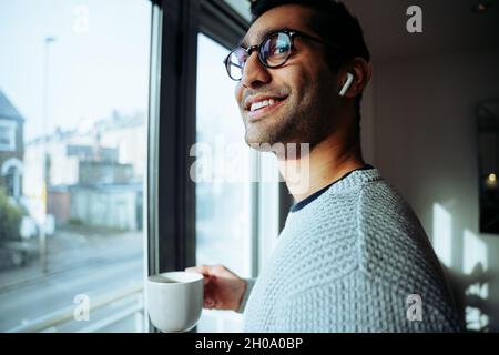 Lächelnder, gemischter Geschäftsmann, der von zu Hause aus arbeitet und morgens mit Kopfhörern einen heißen Kaffee genießt und Musik hört Stockfoto
