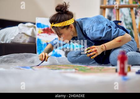 Junge Frau, weibliche Malerin, die konzentriert auf die Malerei schaut, sitzt auf dem Boden in der Werkstatt des Heims Stockfoto