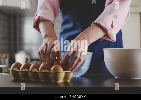 Bäckerin in blauer Schürze, die Eier zum Backen nimmt. Heimelige Kochästhetik. Stockfoto