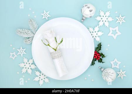 Weihnachtliche Tischdekoration mit weißem Teller, Serviette, Mistel, Baumbaudeko, Stechpalme und Schneeflocken auf blauem Hintergrund. Festliches Design. Stockfoto