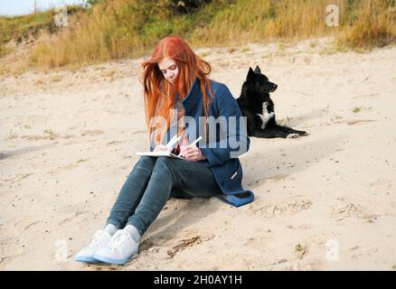 Junge rothaarige Frau zeichnet eine Skizze in einem Skizzenbuch mit Markierungen. Ein schwarzer Hund liegt neben ihr. Erholung mit Haustieren, Sandstrand und Herbst wir Stockfoto