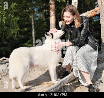 Vorderansicht einer jungen lächelnden Frau, die ihren Hund streichelt, der unter dem Baum in einem Wald sitzt. Spaziergang mit Hund, Liebling, Zweisamkeit und liebevollen Haustieren Stockfoto