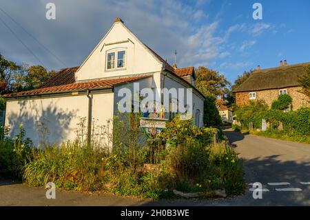 Ein weiß gerendertes traditionelles Backsteinhaus mit dem Dorfschild davor an der Ecke eines steilen Hügels in Happisburgh, Norfolk, England. Stockfoto