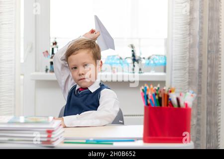 Ein süßer Junge der ersten Klasse in einer Schuluniform zu Hause, während er an seinem Schreibtisch isoliert ist, macht während der Aussparung ein Papierflugzeug. Selektiver Fokus. Hochformat Stockfoto