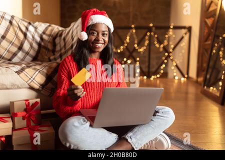 Fröhliche schwarze Frau mit Weihnachtsmütze, Kreditkarte und Laptop, die Weihnachtsgeschenke zu Hause im Internet bestellt Stockfoto