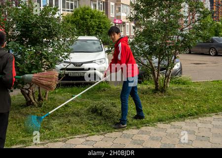 Ein junger asiatischer Hausmeister reckt im Sommer frisch geschnittenes Gras mit einem Gartenreke auf einer Wiese in der Nähe eines Wohnhauses. Stockfoto