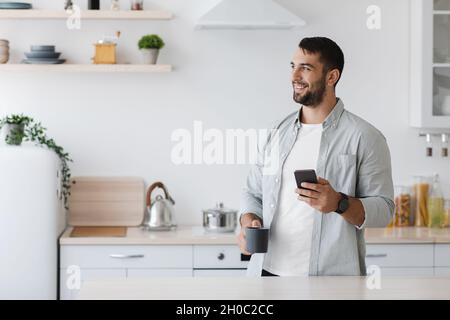 Lächelnder kaukasischer Mann mittleren Alters mit Bart, Smartphone liest Nachrichten und trinkt Kaffee Stockfoto