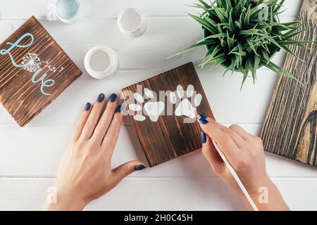 Womans Hand zeichnet Pfoten-Prints auf dekoratives Brett aus verbranntem Holz mit Pinsel aus weißer Farbe. Kreatives DIY Stockfoto