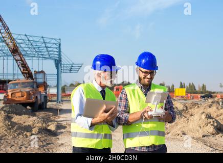 Zwei Ingenieure mit Helmen und Westen, die per Fernbedienung mit der Drohne arbeiten. Technologische Innovationen in der Bauindustrie Stockfoto