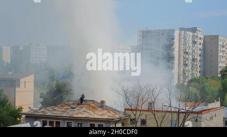 Feuerwehrleute, die auf das Dach des Hauses gehen, um das Feuer aus dem Gebäude in Flammen zu löschen. Feuerwehrmänner der Feuerwehr, deren Ausrüstung versucht, mit Smog und Rauch Flammen aus dem brennenden Haus zu löschen Stockfoto