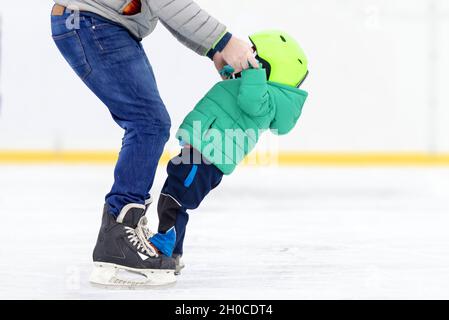 Schwierigkeiten der ersten Schritte auf Schlittschuhe. Kind macht erste Schritte auf Schlittschuhe mit Unterstützung in der Eisbahn. Hintergrund nicht fokussiert. Stockfoto