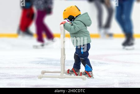 Schwierigkeiten der ersten Schritte auf Schlittschuhe. Kind macht erste Schritte auf Schlittschuhe mit Unterstützung in der Eisbahn. Hintergrund nicht fokussiert. Stockfoto