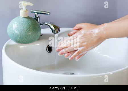 Händewaschen in einem runden weißen Keramikbecken, Seifenblasen zur Hand. Auf dem Waschbecken befindet sich eine grüne Flüssigseifenflasche und Wasser fließt aus dem Wasserhahn Stockfoto