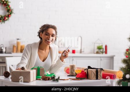 Positive afroamerikanische Frau mit Mobiltelefon in der Nähe von Geschenken, weihnachtsdekor und Tee Stockfoto