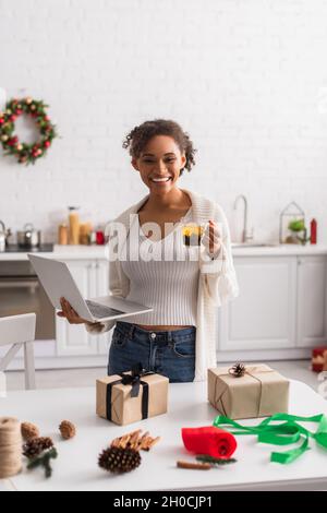 Lächelnde afroamerikanische Frau mit Tee und Laptop in der Nähe von Geschenkboxen und weihnachtsdekor Stockfoto