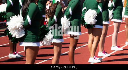 High School Cheerleader stehen auf einer Strecke mit ihren weißen Pom Poms hinter dem Rücken während eines Fußballspiels. Stockfoto