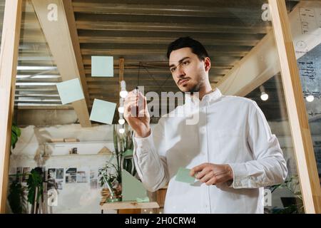Junger bärtiger Manager in einem Hemd, der auf einer klaren Plastiktafel in einem Holzrahmen schreibt. Frontalansicht. Stockfoto