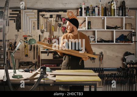 Zimmermann in einer Uhrenkappe mit einem Stück Holz in einer großen Werkstatt. Anzahl der an der Wand hängenden Werkzeuge. Stockfoto