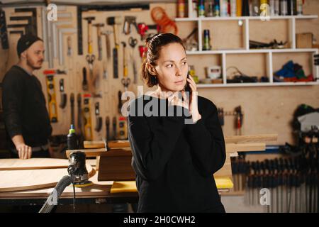 Kundin, die in einer Schreinerei telefoniert. Crew im Hintergrund. Werkzeuge hängen an der Wand. Stockfoto