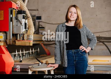 Porträt einer anständigen jungen Arbeiterin in halboffizieller Kleidung, die neben einem großen roten Stück schwerer Maschinerie steht. Sie legte ihre Hand auf ein sto Stockfoto