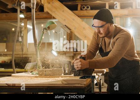 Scharfer Zimmermann in einer Uhrenmütze, der Holz mit einem elektrischen Flugzeug in einer großen Werkstatt rasiert. Anzahl der an der Wand hängenden Werkzeuge. Stockfoto