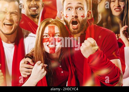 Nahaufnahme eines jungen, emotionalen Mannes und Mädchens, Fußballfans aus England, die ihre Mannschaft mit weißen roten Schals im Stadion anfeuern. Stockfoto